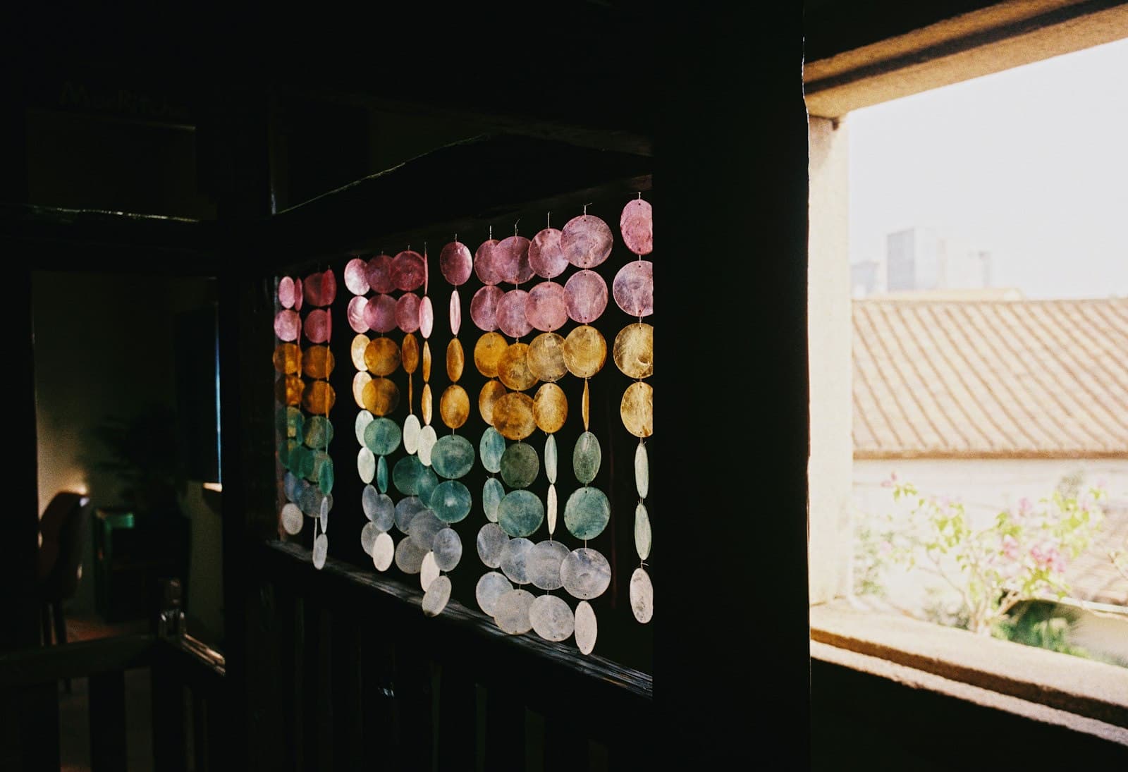 A curtain of translucent dyed shell discs in pink, gold and green hanging beside a sunlit window