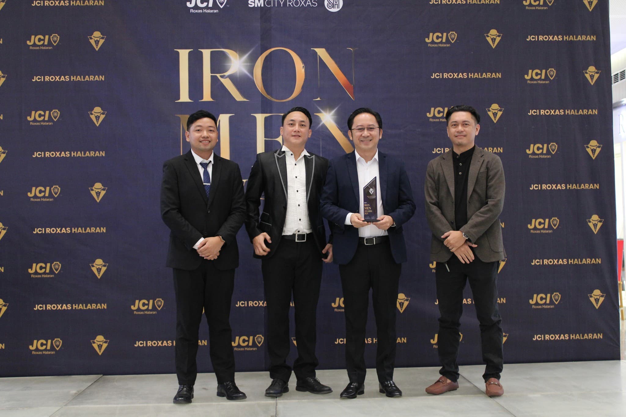 Four men in suits, one holding a trophy, before the JCI Roxas Halaran Iron Men Awards 2025 backdrop